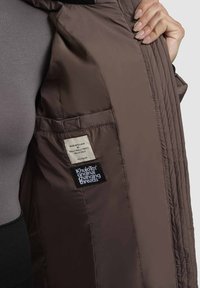 Brauner, gesteppter Jackeninnenraum aus glattem Stoff, mit Etiketten und einer Tasche. Das Design umfasst dezente Nähdetails.