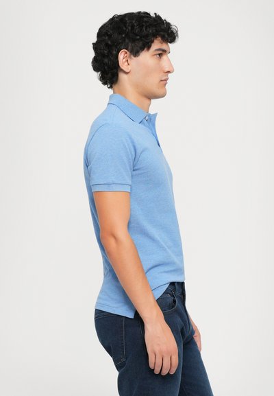 Blauwe polo met korte mouwen en een klassieke kraag, gemaakt van een gestructureerde stof, gedragen met donkerblauwe jeans, zijwaarts getoond.