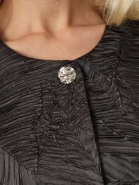 Juodas tekstūruotas drabužis su klostuotu dizainu ir apvaliu metaliniu mygtuku prie kaklo, pabrėžiantis audinio blizgesį ir detales.