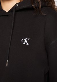 Personne portant un sweat à capuche noir avec le logo blanc "cK" brodé sur la poitrine ; cheveux bruns visibles en haut à droite.