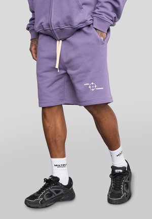 Purple cotton shorts with a soft texture, drawstring waist, and white printed branding on the lower right side. Complemented by black sneakers.