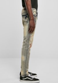 Falmede blå denimjeans med slitte detaljer og malingssprut. Har en slim fit, fem-lommers design og kontraststing.