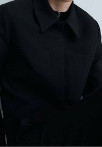 Veste noire ajustée, avec un col pointu, une fermeture éclair et deux poches avant. Texture lisse avec une coupe structurée et un design minimaliste.