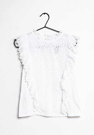 Weiße ärmellose Bluse mit Lochstickerei, gezacktem Rüschenbesatz vorne und an den Schultern, an einem schwarzen Bügel vor weißem Hintergrund hängend.
