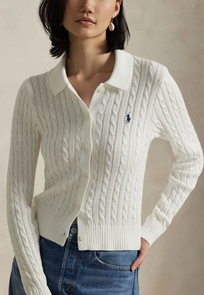 Hvit kabelstrikket cardigan med krage, knappelukking og ribbestrikkede mansjetter. Har en liten blå logo på venstre bryst.