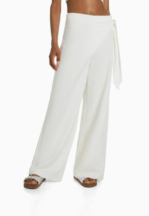 Frau trägt hoch taillierte, weit geschnittene weiße Hosen mit seitlicher Schleife und offenen Sandalen, der Bauchbereich freigelegt.