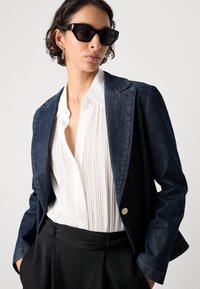 Denim-blazer i mørkeblå med gullhardware, kombinert med en hvit, plissert skjorte og svarte bukser med en skreddersydd passform og strukturert design.