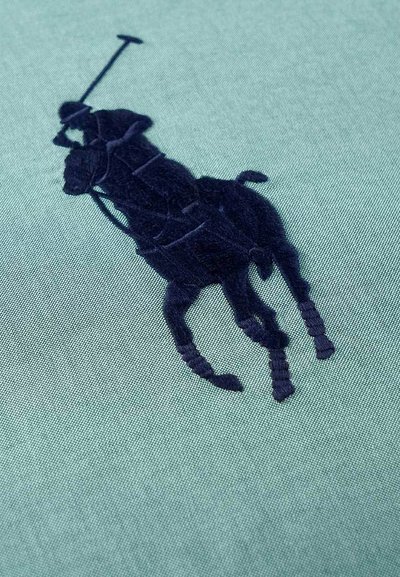 Πράσινο βαμβακερό ύφασμα με κέντημα ενός παίκτη πόλο πάνω σε άλογο σε σκούρο ναυτικό χρώμα, με λεπτομέρεια σε υφή και λεία επιφάνεια.