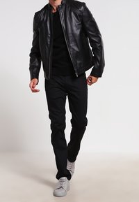 Czarna skórzana kurtka z stojącym kołnierzem, noszona na czarnym swetrze, w połączeniu z czarnymi spodniami i jasnoszarymi trampkami.