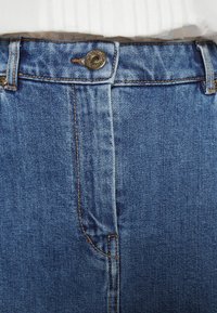 Gros plan sur la ceinture d’un jean en denim bleu avec un bouton en métal et des passants de ceinture visibles, porté avec une chemise blanche rentrée.