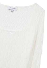 Blusa bianca in pizzo con scollatura tonda, delicato motivo floreale e spalline sottili. Presenta un piccolo fiocco in raso sul petto.