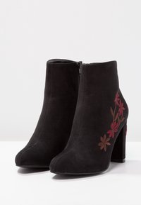 Bottes cheville en daim noir avec talons carrés, fermeture éclair latérale, et broderie florale rouge et marron sur les côtés externes.