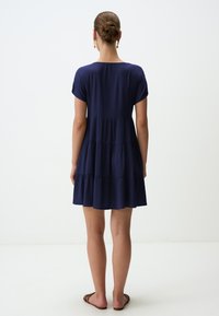 Kurzkleid in Marineblau mit Rundhalsausschnitt, kurzen Ärmeln und schichtweisen Rüschen. Glatte Textur, knielang, kombiniert mit flachen Sandalen.