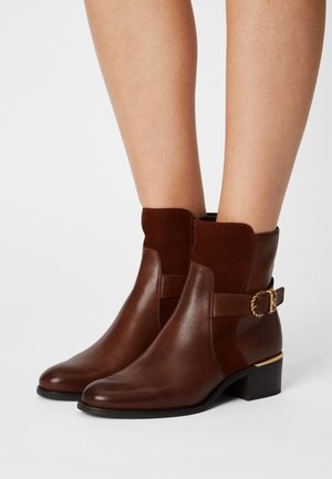 Botas de tobillo de cuero marrón con tacones de bloque, paneles de ante en el tobillo y correas decorativas con hebillas doradas, usadas sobre piernas desnudas.