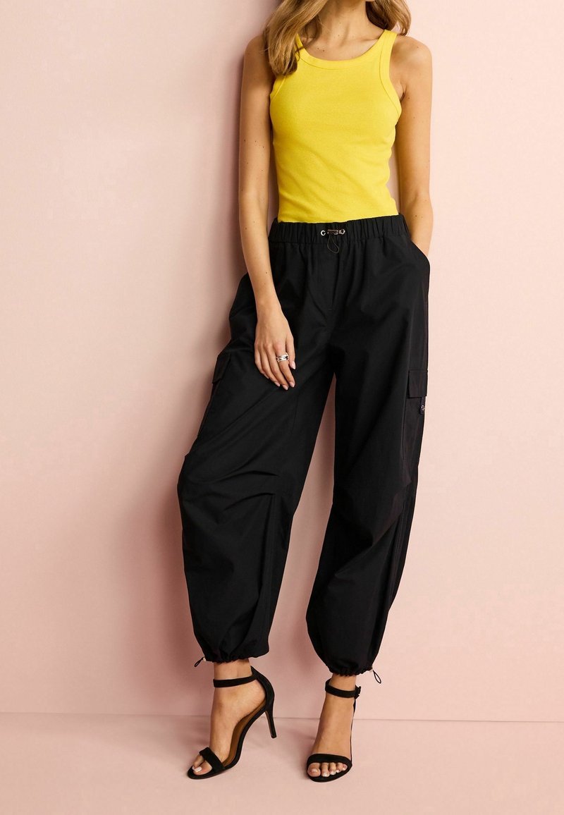 Un pantalon cargo noir avec des poignets élastiques, associé à un débardeur jaune ajusté et des sandales à talons noirs avec des lanières. Les pantalons disposent de poches latérales.
