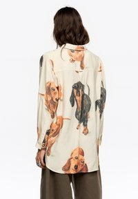 Beżowa koszula z długim rękawem, z nadrukiem przedstawiającym jamniki w różnych pozach. Wykonana z lekkiego materiału, z klasycznym kołnierzykiem.