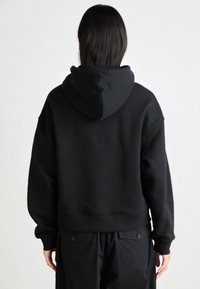 Personne aux longs cheveux foncés portant un sweat à capuche noir et un pantalon noir, debout face à un fond gris clair uni.