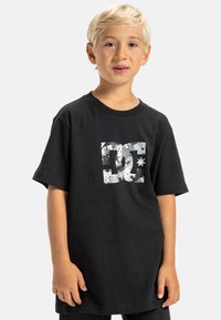 Camiseta de algodón negra con un logo de DC en gris y blanco desgastado que incluye una estrella, corte relajado, mangas cortas y cuello redondo.