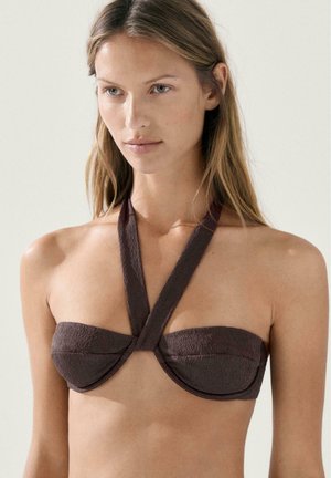 Mladá žena s dlouhými vlasy, která má na sobě texturovaný tmavě hnědý halterový bikiny top na jednobarevném světlém pozadí.