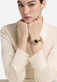 Moteris kreminės spalvos palaidinėje, laikanti smakrą ant kumščių, kairėje rankoje dėvi auksinį laikrodį su juodu ciferblatu ir odiniu dirželiu.