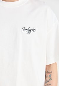 Camiseta de algodón blanca con un pequeño logo bordado en negro de "Carhartt WIP" en el área del pecho izquierdo. Mangas cortas y corte clásico.