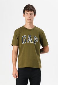Joven con cabello corto castaño que lleva una camiseta deportiva GAP de color verde oliva y pantalones negros, de pie contra un fondo blanco liso.