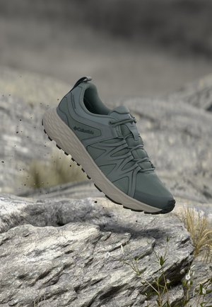 Scarpa da trekking impermeabile Columbia grigia sospesa sopra un terreno roccioso con piccole piante e terra sparsa.