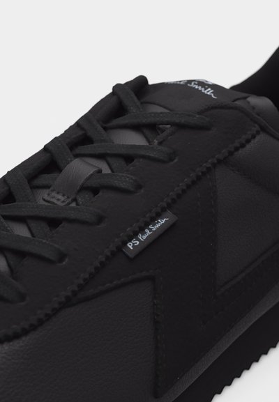 Κοντινή λήψη μαύρου αθλητικού παπουτσιού με υφασμάτινο σχέδιο σε σχήμα "V", μαύρα κορδόνια και μικρή υφασμάτινη ετικέτα "PS Paul Smith" στο πλάι.
