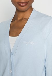 Primer plano de una persona que lleva un cárdigan abotonado de color azul claro sobre una blusa blanca, mostrando el nombre de la marca bordado en el pecho.