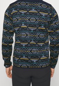 Námořnický pletený svetr s geometrickými vzory v modré, žluté a šedé barvě. Má dlouhé rukávy a kulatý výstřih, s texturovaným povrchem.