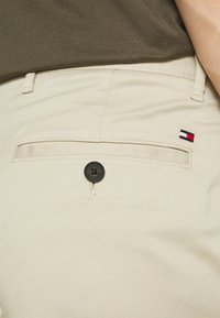Khaki kalhoty s přední kapsou, která má černý knoflík a malý logo Tommy Hilfiger v červených, bílých a modrých pruzích na zadní straně.