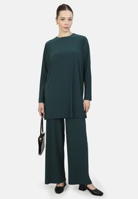 Tunik berlipit hijau tua dan celana lebar, terbuat dari kain halus. Dipasangkan dengan tas tangan hitam dan sepatu hitam berhak rendah.