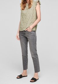 Haut sans manches vert olive avec des accents de volants aux épaules, motif floral texturé ; associé à un jean gris droit et des sandales plates noires.