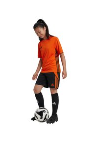 Camiseta deportiva naranja de manga corta con rayas negras, combinada con pantalones cortos negros con detalles naranjas y calcetas negras, de pie junto a un balón de fútbol.