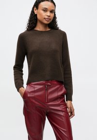 Donna con i capelli ricci che indossa un maglione a maglia marrone scuro e pantaloni in pelle rossa a vita alta con cerniera frontale e tasche, in piedi su uno sfondo bianco.