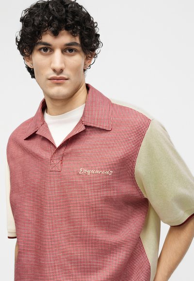 Jeune homme aux cheveux bouclés portant un polo à manches courtes rouge et beige avec un petit logo sur la poitrine, sur fond blanc.