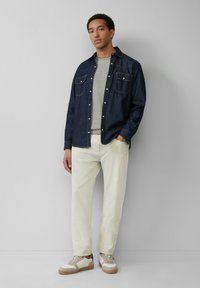 Jonge man in een donkerdenimjas over een grijs shirt, lichte beige broek en beige sneakers met bruine zolen, staande tegen een grijze muur.