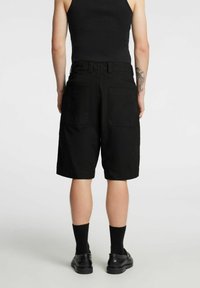 Pantalones cortos de algodón negros con corte recto, dos bolsillos traseros y cintura con trabillas para cinturón. Se usan con calcetines y zapatos negros. Diseño sencillo.