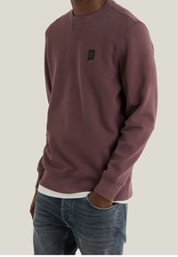 Felpa viola con collo rotondo e polsini a costine, caratterizzata da un piccolo patch con logo nero sul petto; indossata sopra una camicia bianca.