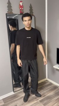 Schwarzes T-Shirt mit "PEGADOR"-Logo, kombiniert mit schwarzer Hose, stehend vor einem Spiegel. Holzboden und schlichte Dekoration im Hintergrund.
