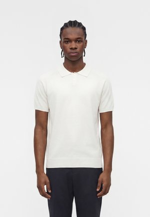 Άντρας με πλεξούδες, φοράει λευκό πόλο με κοντά μανίκια και σκούρο παντελόνι, στέκεται μπροστά σε ένα απλό φωτεινό φόντο.