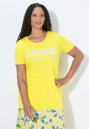Kvinde med langt krøllet hår iført en gul "FRESH & Juicy" skjorte og et skørt med citron- og grønne bladmønstre.