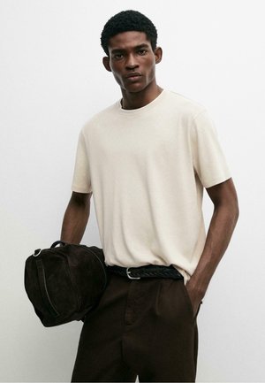 Jonge man met een beige shirt met korte mouwen en donkere broek, houdt een zwarte tas in één hand en de andere hand in zijn zak.