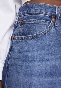 Siniset farkut oranssilla ompeleella, viisipokkisuus ja korkeavyötäröinen malli. Sileä pinta, jossa on hieman haalistumaa kankaassa.