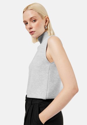 Blonde Frau mit ärmellosem grauem Rollkragenpullover und schwarzer Hose mit hoher Taille, posiert mit einer Hand in der Tasche, neutraler Hintergrund.