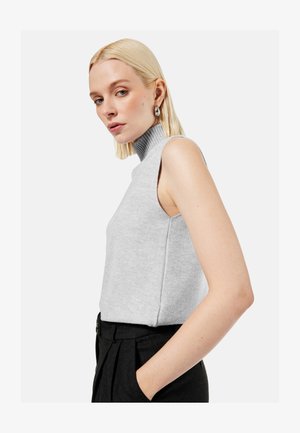 Blonde Frau mit ärmellosem grauem Rollkragenpullover und schwarzer Hose mit hoher Taille, posiert mit einer Hand in der Tasche, neutraler Hintergrund.