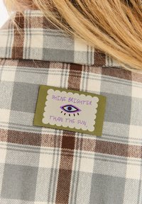 Naszywka z niebieskim okiem i napisem „Błyszcz jaśniej niż słońce” na plecach brązowo-beżowo-szarej koszuli w kratę, z widocznymi blond włosami.