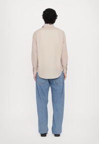 Camicia a maniche lunghe di colore beige chiaro con una texture liscia e orlo curvo, abbinata a jeans larghi di colore blu chiaro. Scarpe nere visibili.