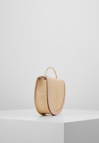 Mała beżowa torba pleciona z zaokrąglonymi krawędziami i złotym uchwytem na górze, stojąca w pozycji pionowej na białym piedestale na szarym tle.