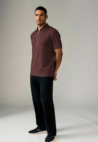 Camisa polo burdeos con un patrón de zigzag texturizado, con cremallera frontal, combinada con pantalones negros y zapatillas oscuras frente a un fondo neutro.